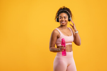 A woman smiles while holding a pink water bottle and wearing headphones. She's dressed in fitted...