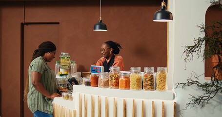 Local vendor putting merchandise on scale at checkout, ensuring the best pricing value for regular client at grocery store. African american retailer serving buyer, eco friendly. Handheld shot.