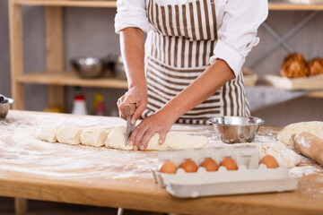 Close-up of skilled baker hands cutting soft dough with scraper on floured wooden worktable, filled...