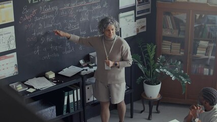 Middle-aged female teacher conducting lesson in classroom, standing in front of blackboard filled with handwritten English grammar notes, explaining concepts to students. High angle view