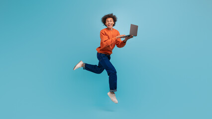A young man with curly hair energetically leaps in an airy setting, holding a laptop while...