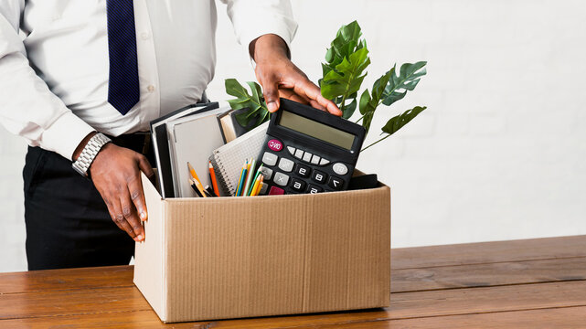 Unrecognizable black guy collecting his office stuff into carton box. Closeup of African American man losing job, leaving workplace with his belongings. Economic crisis and unemployment