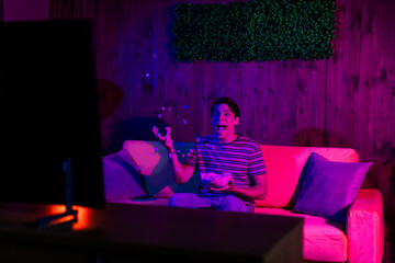 Young man laughing and throwing popcorn while watching TV in a vibrant and cozy living room