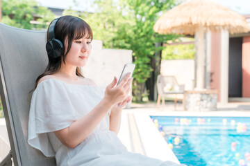 プールサイドのリクライニングチェアに座り音楽を聴く若いアジア人女性
