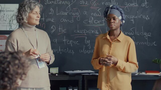 Young Black female student wearing headscarf standing at chalkboard, reciting English lesson in front of middle-aged teacher and classmates