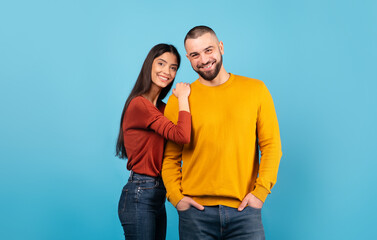 Naklejka premium A millennial couple stands close together against a bright blue backdrop. The woman smiles warmly, while the man displays a charming grin, embodying happiness and connection in a candid moment.