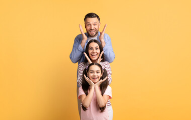 Parents and their teenage daughter stand together against a bright yellow background, happily posing in a playful manner that shows their love and affection for each other.