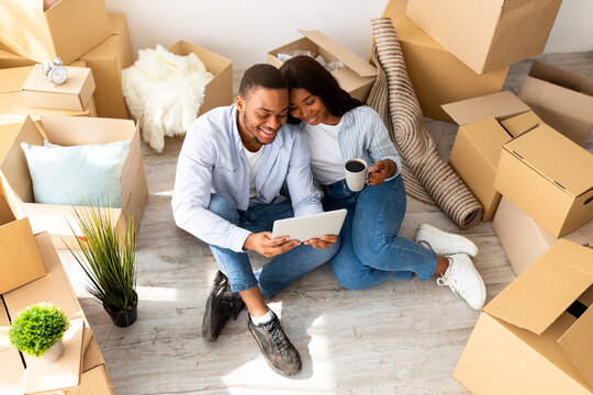 Happy african american family sitting on floor in new apartment with digital tablet among boxes with belongings, buying furniture for house or working on design together, above view