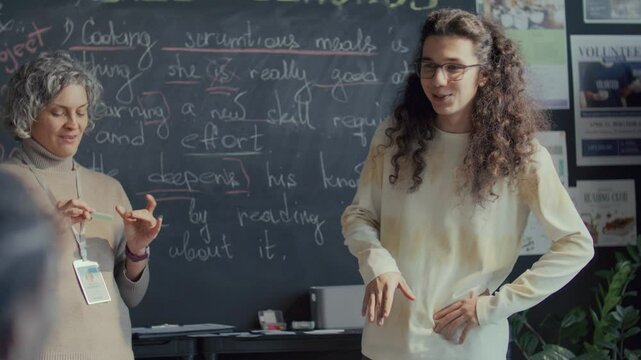 Young gen Z student with long curly hair standing in front of blackboard, smiling and talking to teacher and classmates during English lesson in high school