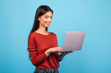 A young brunette woman stands in front of a vivid blue backdrop, happily typing on her laptop. She exhibits a relaxed and content demeanor, embodying a modern lifestyle.