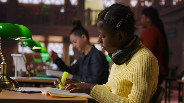 Black preteen pupil underlining key words and phrases for the learning session at the library, creating a summary to retain information easier. Focused scholar studying. Camera B.