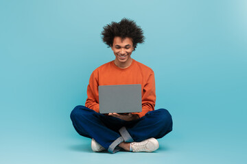 A young African American man with curly hair sits on the floor, smiling while using a laptop. He wears a casual outfit and the background is a solid light blue color.