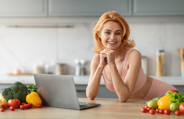 A woman with blonde hair smiles while leaning on her hands, surrounded by fresh vegetables and a laptop, indicating a healthy cooking session or meal planning at home.