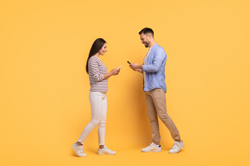 A family couple stands close together against a vibrant yellow backdrop, engaging with their smartphones. They share smiles and laughter, reflecting their affectionate bond and enjoyment.