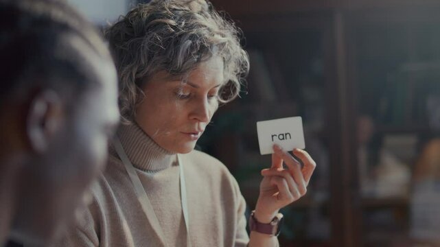 Female English teacher with curly grey hair demonstrating flashcard labeled Ran, explaining verb past tense to students in classroom during lesson