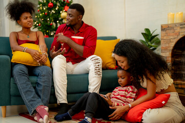 A cheerful family gathers around the Christmas tree, sharing a moment of joy as a man presents a red gift box to a child. while family members relax on the sofa, enjoying the warmth of the season.