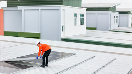 technician in hard hat and safety gear inspects solar panels on a metal rooftop, holding a tool....