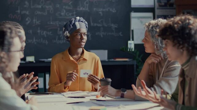 African American female student holding flashcard labeled Ate and discussing it with teacher and diverse classmates, learning new words during English lesson