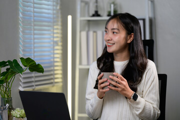 Fototapeta premium Young, beautiful asian woman savoring a warm cup of coffee while enjoying a well-deserved break from working on her laptop in a cozy home office filled with natural light