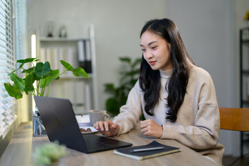 Young asian businesswoman is working from home, using her laptop at a desk with plants, coffee and...