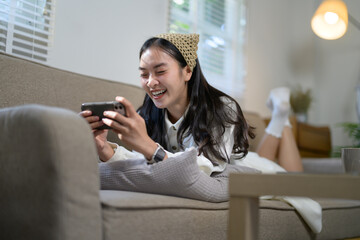 Cheerful young woman lounging on a cozy sofa, using her smartphone to play a mobile game and fully...