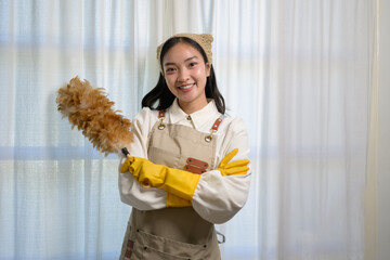 Young asian housekeeper wearing an apron and yellow gloves, smiling brightly while holding a feather duster, ready to tackle house cleaning tasks with enthusiasm