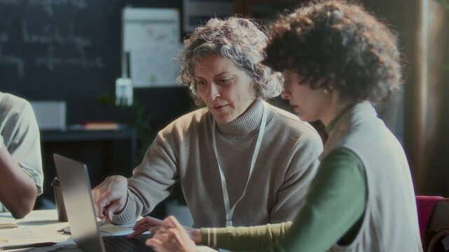 Middle-aged teacher and young female student looking at laptop screen and discussing information at table in classroom during English lesson. Rack focus