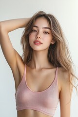 Fototapeta premium Elegant young Asian woman in soft pink tank top lifting hand showcasing clean underarms on white background
