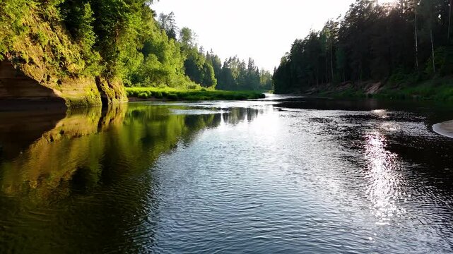 Drone pullback past serene riverside clifs with sun glowing through trees, Gauja River Ligatne Latvia