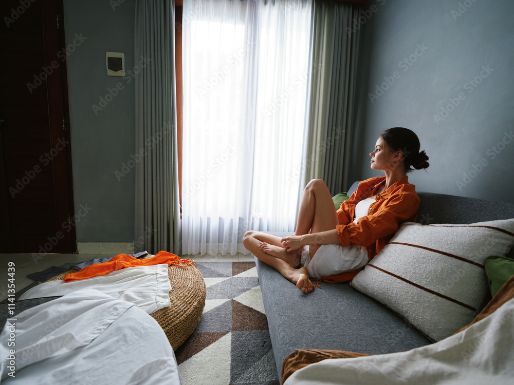 Wall mural A serene young woman in casual attire sitting by a window, enjoying a moment of solitude in a cozy modern room with a soft color palette