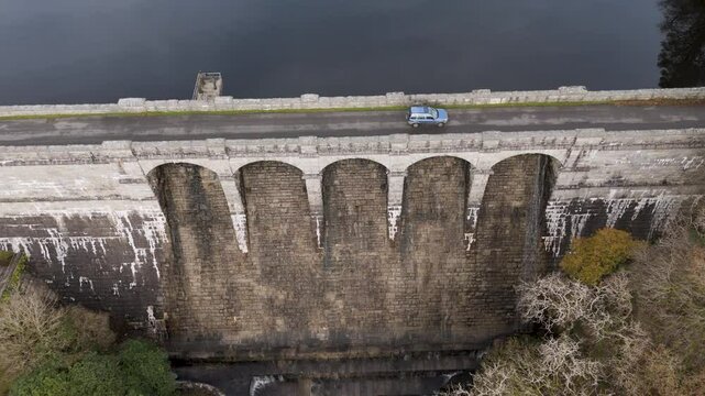 High-angle aerial of a blue off-road vehicle crossing a time-worn stone bridge in a rural setting, evoking adventure, exploration, and the thrill of remote travel