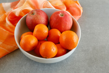A white bowl filled with fresh red apples and vibrant orange tangerines sits on a gray countertop. An orange fabric drapes in the background, adding a warm and colorful contrast.