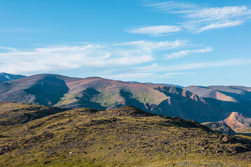 Scenic alpine landscape with sunlit big green hill against high vivid rocky mountain range in bright sun under clouds in blue sky. Colorful large mountains with light and shadows on just before sunset