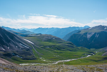 Aerial top view to sunlit green alpine valley with creek among big hills and large rocky mountains silhouettes in bright sun under clouds in blue sky. High snowy mountain range far away in sunny day.