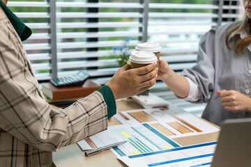 Two people are holding coffee cups and shaking hands. The scene is set in an office with a desk and a window