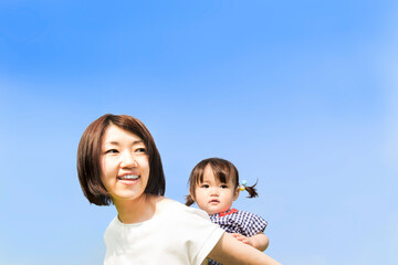 Fototapeta premium 青空を背景に女の子の幼児を背負い育児をする若いお母さん。育児,家族,愛情のイメージ
