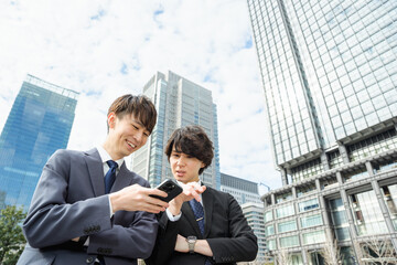 高層ビルに囲まれた都会でスマートフォンを見て笑顔で会話するスーツ姿の20代?30代の2人の男性