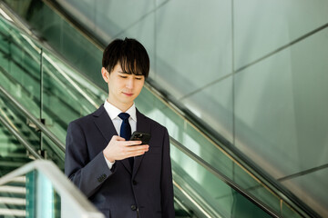 移動中にエスカレーターに乗りながらスマートフォンを操作するスーツ姿の20代の日本人男性