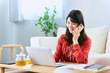 Obraz premium 自宅リビングでのテレワークやパソコンでのオンライン学習や勉強中にスマートフォンの電話で通話をする女性