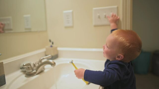 red-haired toddler in blue shirt holds toothbrush while reaching up to toggle light switch in bathroom, showcasing curiosity and independence.Toddler reaching for light switch in bathroom