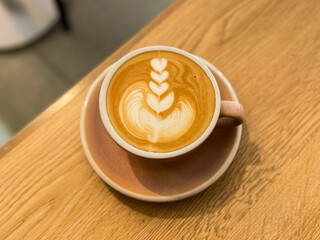 A cappuccino with intricate latte art sits in a pink cup on a wooden table.