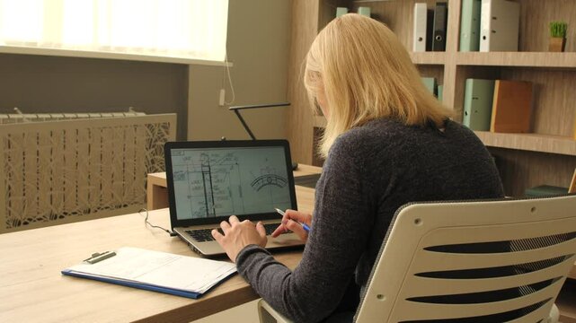Rear view of focused professional middle aged female engineer creating construction design , analyzing project sketch using laptop pc while working on blueprint at office.