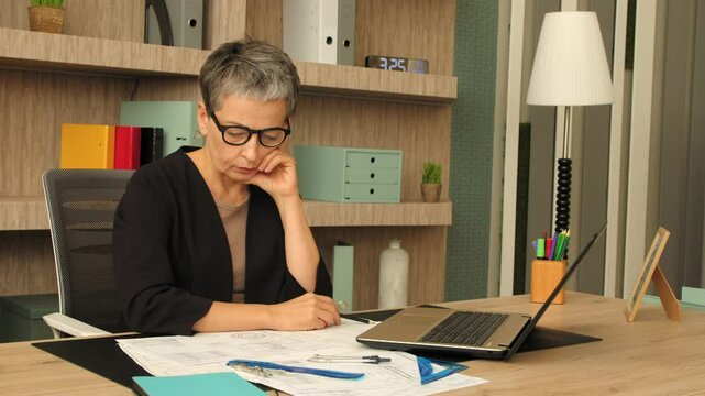 Thoughtful charming middle aged female architect in casual clothes sitting at desk, pondering over house design and drawing sketch with drafting tools while working on project at office .