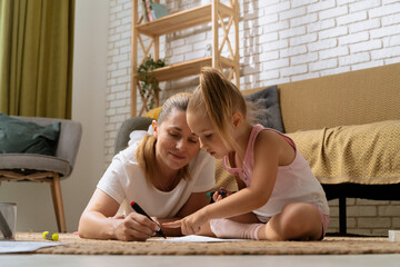 Young mother and daughter sitting on the floor at home spending time together, mom and girl drawing pictures with colorful pens, close shot.