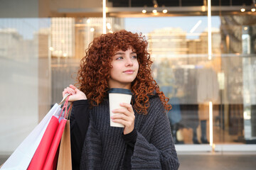 Young woman holding shopping bags and drinking coffee near a mall