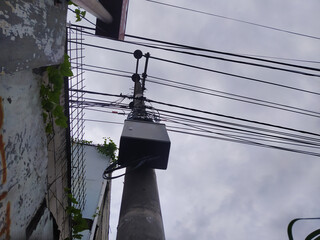 A concrete utility pole with tangled electrical wires, surrounded by walls and overcast sky above