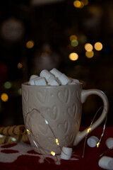 a cup of hot chocolate with marshmallow on the christmas tree with lights background.