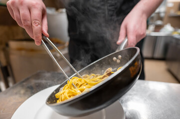 A chef skillfully tosses steaming pasta in a pan using tongs. The scene captures the art of cooking in a professional kitchen, showcasing culinary expertise and delicious food preparation.