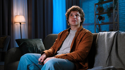 Young man spending evening at home sitting on sofa in living room, watching tv, evening with movie or news program, positive smiling expression.