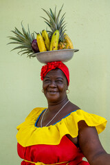 A Palenquera Woman Wearing A Bright Traditional Dress With A Fruit Basket On Her Head, Smiling Against A Green Wall.